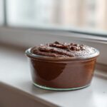 A serving of rich Vegan Chocolate Avocado Mousse in a small glass jar sitting on a white windowsill.