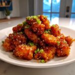 A mound of crispy, glazed Air Fryer Orange Chicken pieces garnished with sesame seeds and sliced green onions on a white plate.