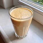 A tall glass filled with a thick, creamy Banana Peanut Butter Smoothie sitting on a windowsill.