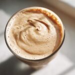 Overhead close-up of the thick, creamy, frothy top of a Banana Peanut Butter Smoothie in a glass.