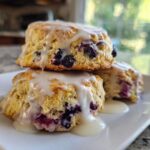 Three freshly baked Blueberry Lemon Scones stacked on a white plate, drizzled heavily with lemon glaze.