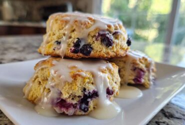 Three freshly baked Blueberry Lemon Scones stacked on a white plate, drizzled heavily with lemon glaze.