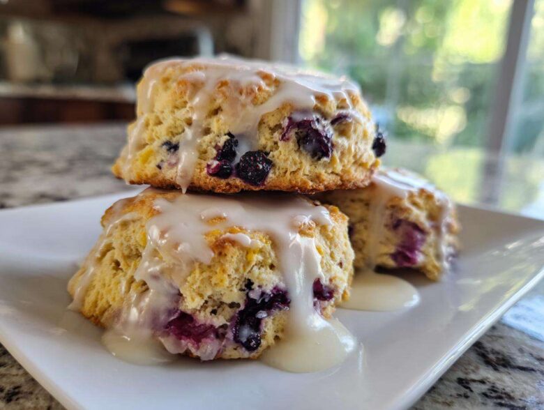 Three freshly baked Blueberry Lemon Scones stacked on a white plate, drizzled heavily with lemon glaze.