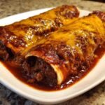 Two Cheesy Beef Enchiladas smothered in rich red sauce and melted cheddar cheese on a white plate.