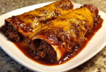 Two Cheesy Beef Enchiladas smothered in rich red sauce and melted cheddar cheese on a white plate.