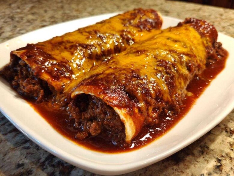 Two Cheesy Beef Enchiladas smothered in rich red sauce and melted cheddar cheese on a white plate.