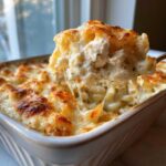 A spoonful of Cheesy Chicken Alfredo Pasta Bake showing a dramatic cheese pull over the baked casserole dish.