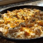 Close-up of Cheesy Chicken and Rice Skillet topped with melted cheddar cheese and fresh parsley in a black pan.