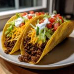 Two hard-shell Cheesy Ground Beef Tacos filled with seasoned meat, shredded cheese, lettuce, tomato, and sour cream.