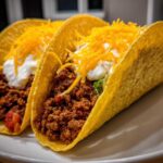 Two crispy shell Cheesy Ground Beef Tacos filled with seasoned meat, sour cream, and shredded cheddar cheese.