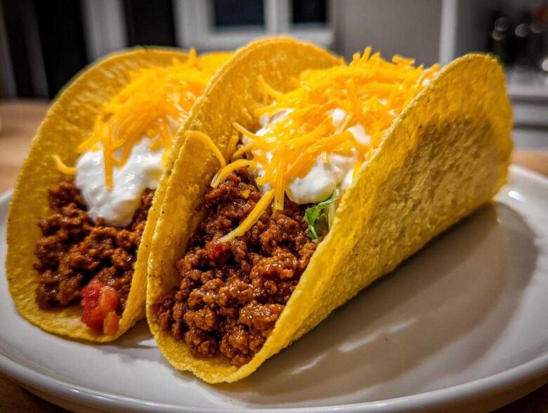 Two crispy shell Cheesy Ground Beef Tacos filled with seasoned meat, sour cream, and shredded cheddar cheese.