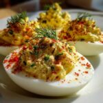 Four halves of Classic Deviled Eggs with Dill Pickle Relish, topped with paprika and fresh dill, served on a white plate.