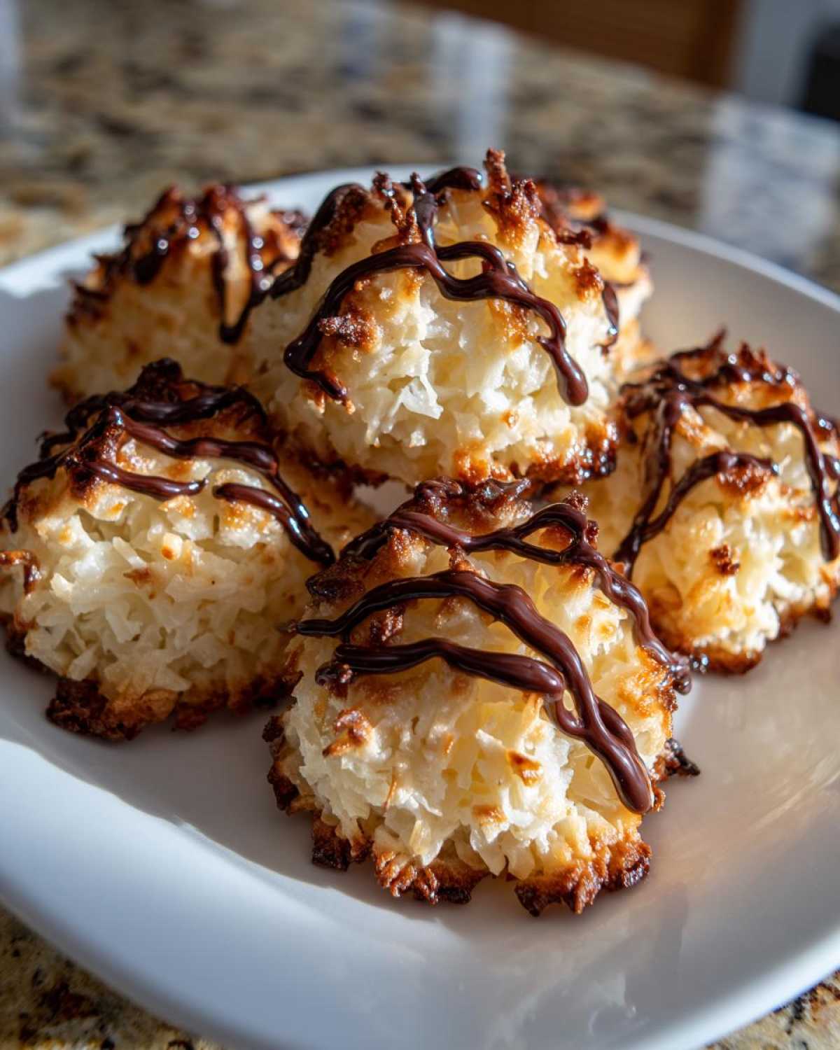 Close-up of golden-brown Coconut Macaroons with Dark Chocolate Drizzle stacked on a white plate.