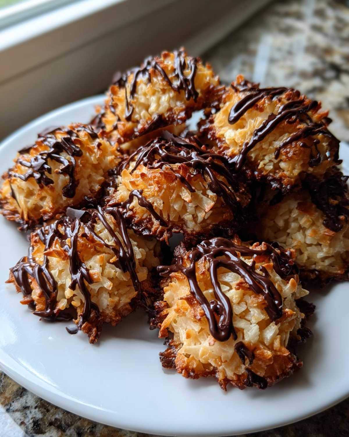 A pile of golden-brown Coconut Macaroons with Dark Chocolate Drizzle stacked on a white plate.