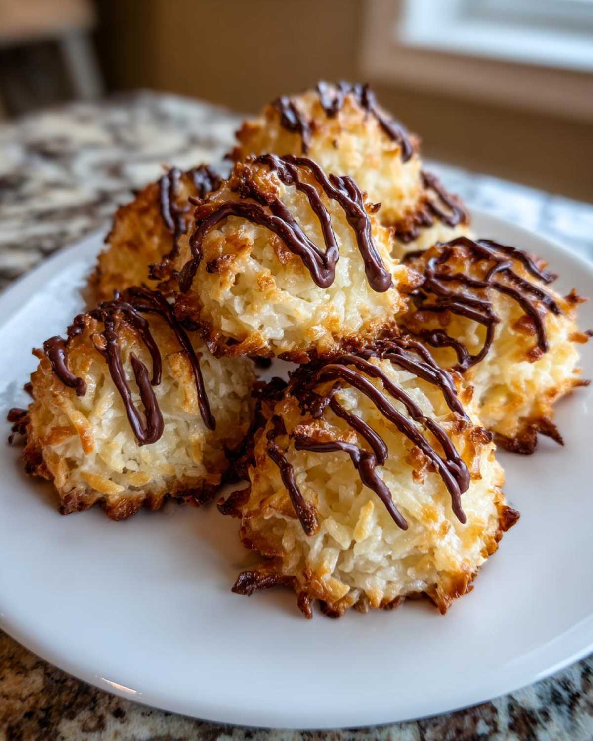 A pile of golden brown Coconut Macaroons with Dark Chocolate Drizzle stacked on a white plate.
