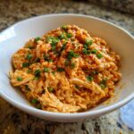 A close-up of a white bowl filled with rich, orange-hued Creamy Cajun Chicken and Rice Skillet, topped with chopped green onions.