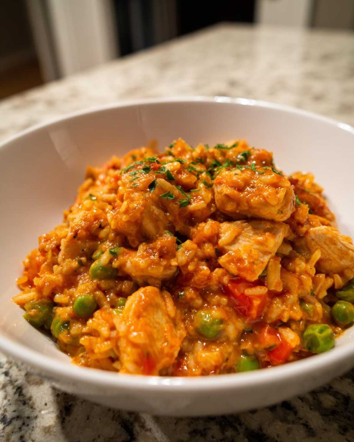 Close-up of a white bowl filled with rich, orange-hued Creamy Cajun Chicken and Rice Skillet, featuring chicken pieces and green peas.