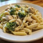 A close-up of a white bowl filled with creamy chicken and broccoli pasta dinner, featuring penne pasta and chunks of chicken.