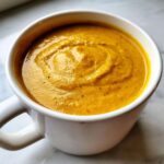 A close-up, top-down view of thick, vibrant orange Creamy Coconut Lentil Soup swirled inside a white mug.