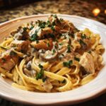 A close-up of fettuccine pasta topped with chunks of chicken, sliced mushrooms, and rich creamy mushroom chicken pasta with garlic sauce, garnished with parsley.