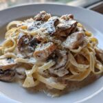 A close-up of fettuccine pasta tossed in a creamy sauce with chicken pieces, sliced mushrooms, and grated Parmesan cheese.