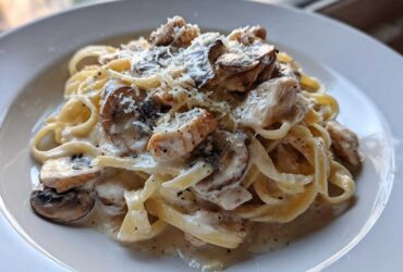 A close-up of fettuccine pasta tossed in a creamy sauce with chicken pieces, sliced mushrooms, and grated Parmesan cheese.