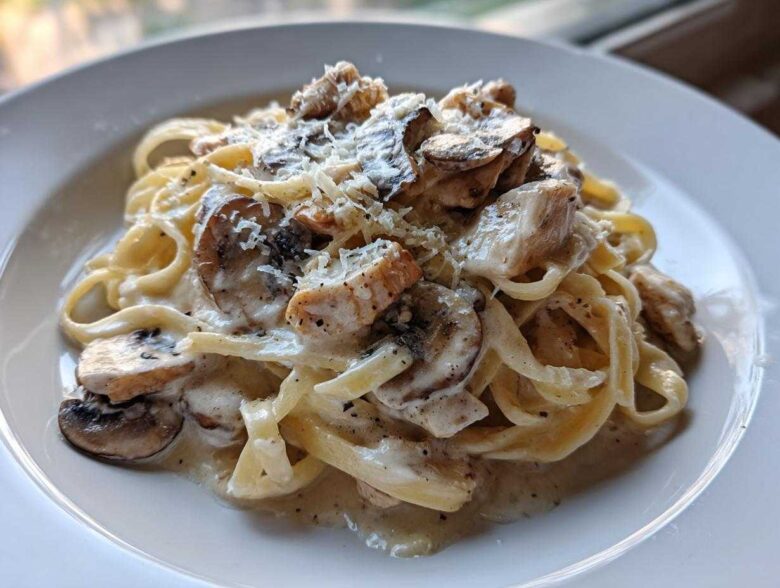 A close-up of fettuccine pasta tossed in a creamy sauce with chicken pieces, sliced mushrooms, and grated Parmesan cheese.
