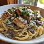 A close-up of fettuccine pasta topped with seared chicken pieces, sliced mushrooms, and a rich, creamy garlic sauce, garnished with parsley.