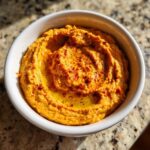 A close-up, overhead shot of creamy Roasted Red Pepper Hummus in a white bowl, drizzled with olive oil and sprinkled with paprika.
