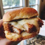 A hand holds a delicious Crispy Chicken Burger with Melty Cheese, showing the golden bun and thick, juicy fried chicken patty.