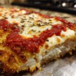 A close-up cross-section of Crispy Chicken Parmesan with melty cheese, bright red marinara sauce, and herbs.