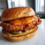 A close-up of a delicious Crispy Chicken Sandwich with Spicy Honey Sauce dripping over the fried fillet and pickles.