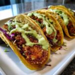 A row of crispy chicken tacos topped with vibrant green avocado cream sauce and purple cabbage slaw.