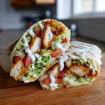 A Crispy Chicken Wrap with Ranch Sauce cut in half, showing crispy chicken, lettuce, tomatoes, and a generous ranch drizzle.
