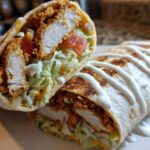 Cross-section of a Crispy Chicken Wrap with Ranch Sauce, showing fried chicken, lettuce, tomato, and a drizzle of white sauce.