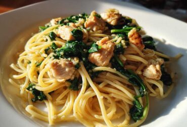 A close-up of a white plate filled with Garlic Butter Pasta with Chicken and Spinach, glistening under natural light.