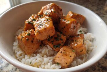 Close-up of glossy Garlic Butter Salmon Bites piled high on a bed of fluffy white rice in a white bowl.