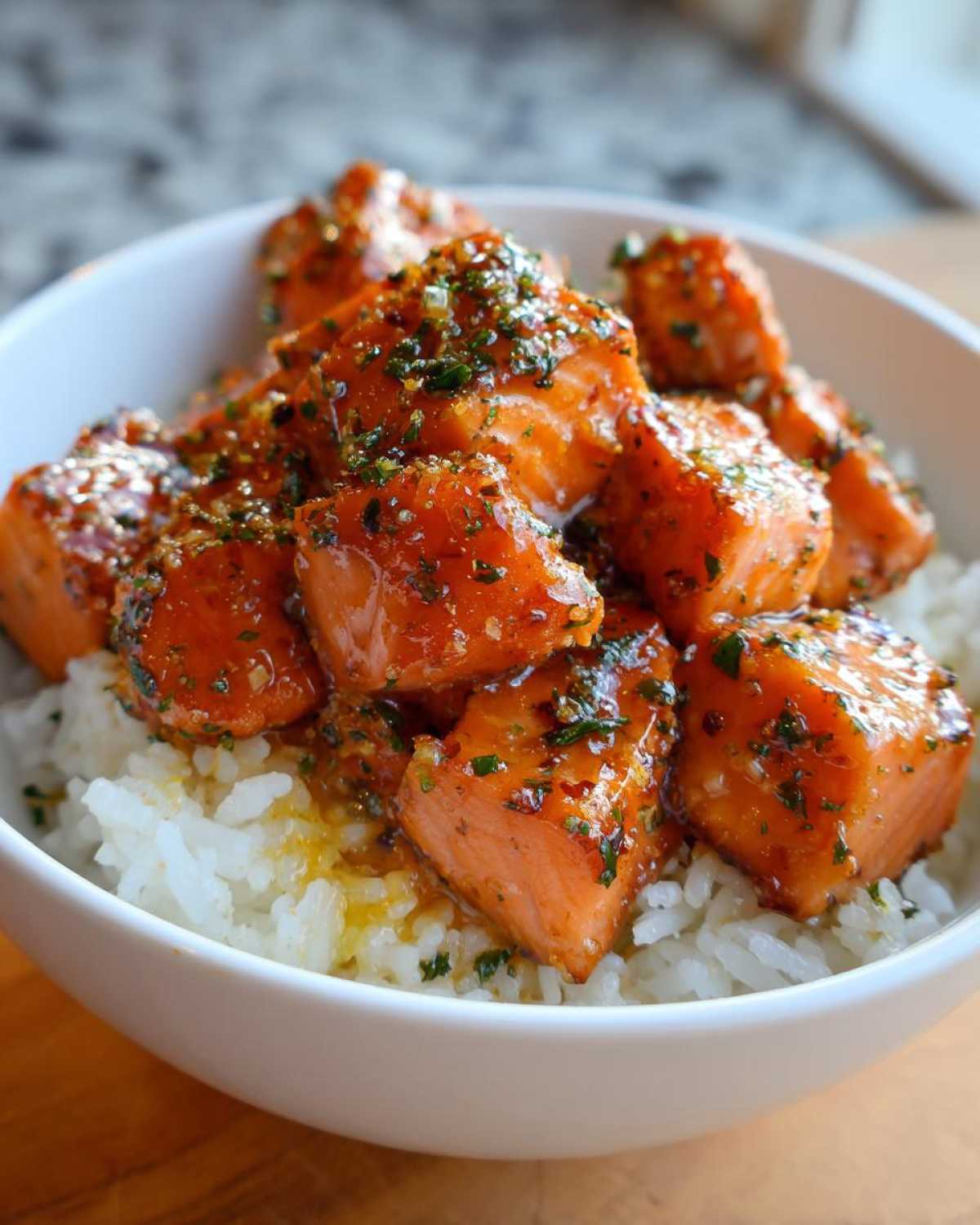 Close-up of glossy Garlic Butter Salmon Bites piled high over a bed of fluffy white rice in a white bowl.