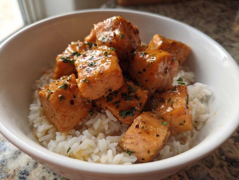 Close-up of glossy Garlic Butter Salmon Bites piled high on a bed of fluffy white rice in a white bowl.