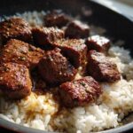 Close-up of seared steak bites coated in garlic butter sauce served over white rice in a black skillet.