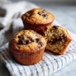 Three freshly baked Gluten-Free Banana Chocolate Chip Muffins, one of which is broken open to show the moist interior.