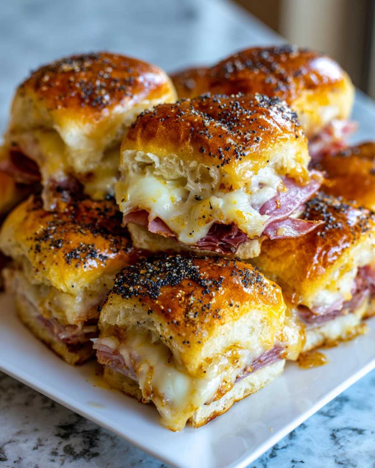 Close-up of gooey Hawaiian Roll Ham and Cheese Sliders stacked on a white plate, topped with poppy seeds.