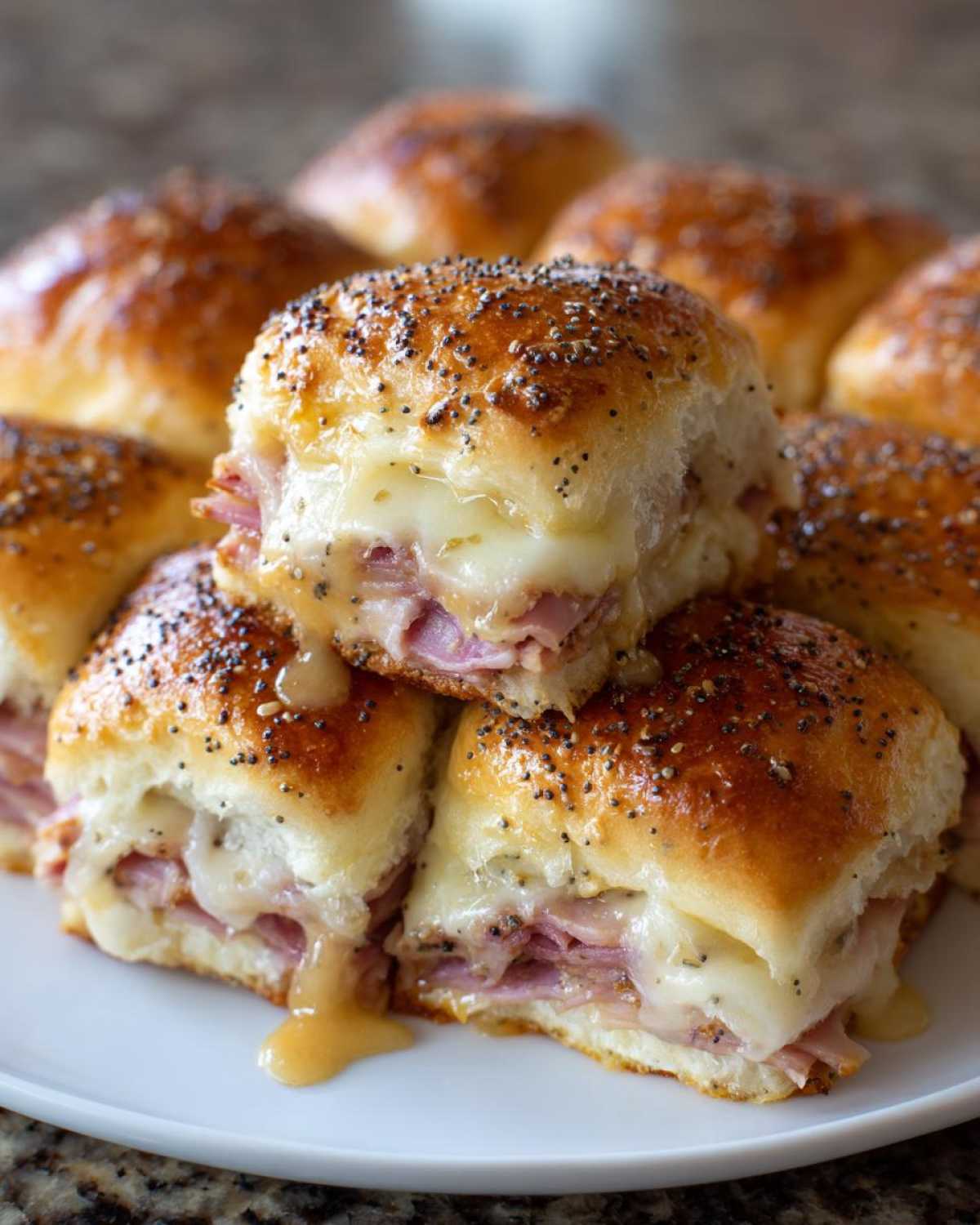 Close-up of melted Hawaiian Roll Ham and Cheese Sliders stacked on a white plate, drizzled with glaze and topped with poppy seeds.
