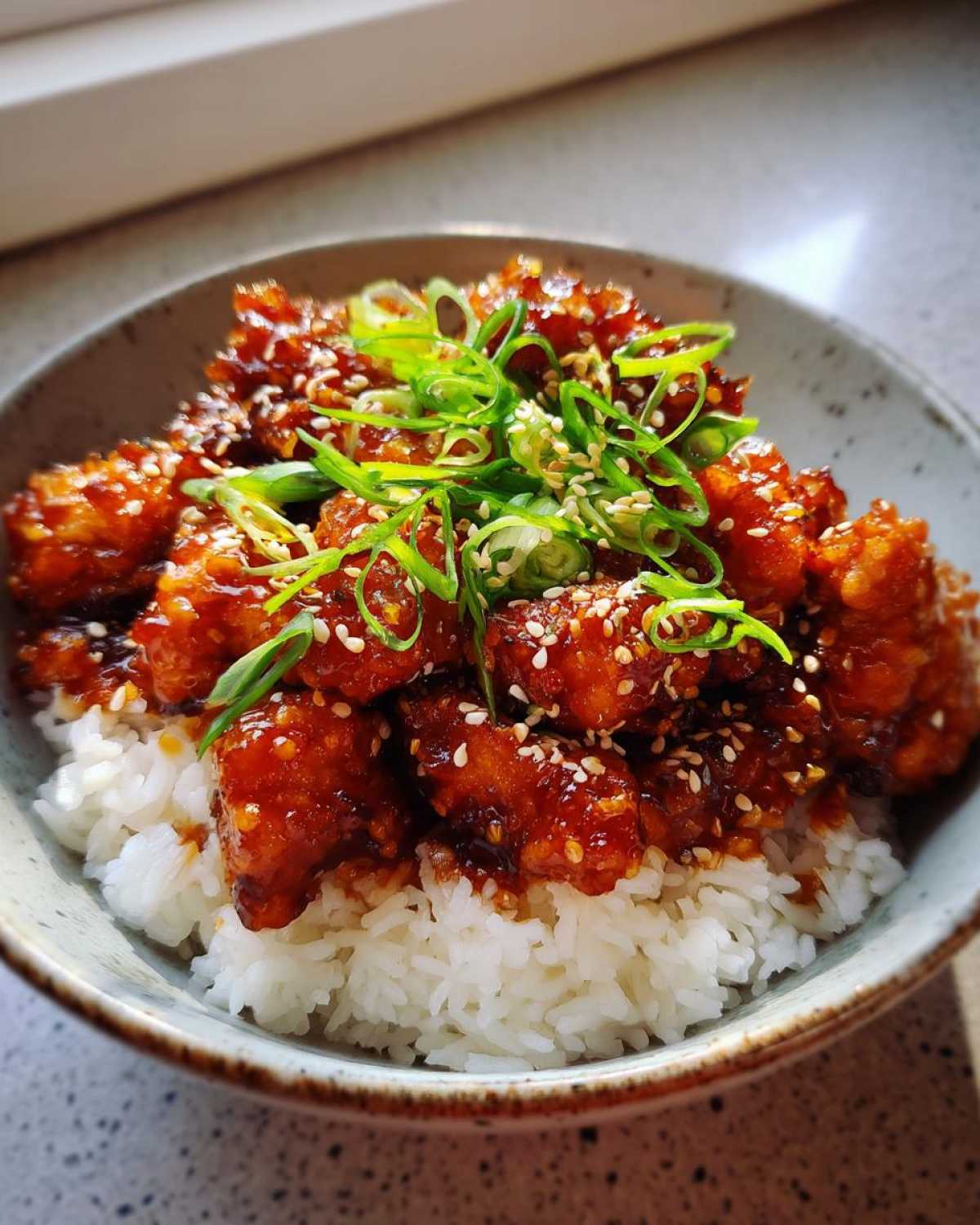 Close-up of a Honey Garlic Chicken Bowl with rice, topped with sticky glazed chicken, sesame seeds, and fresh scallions.