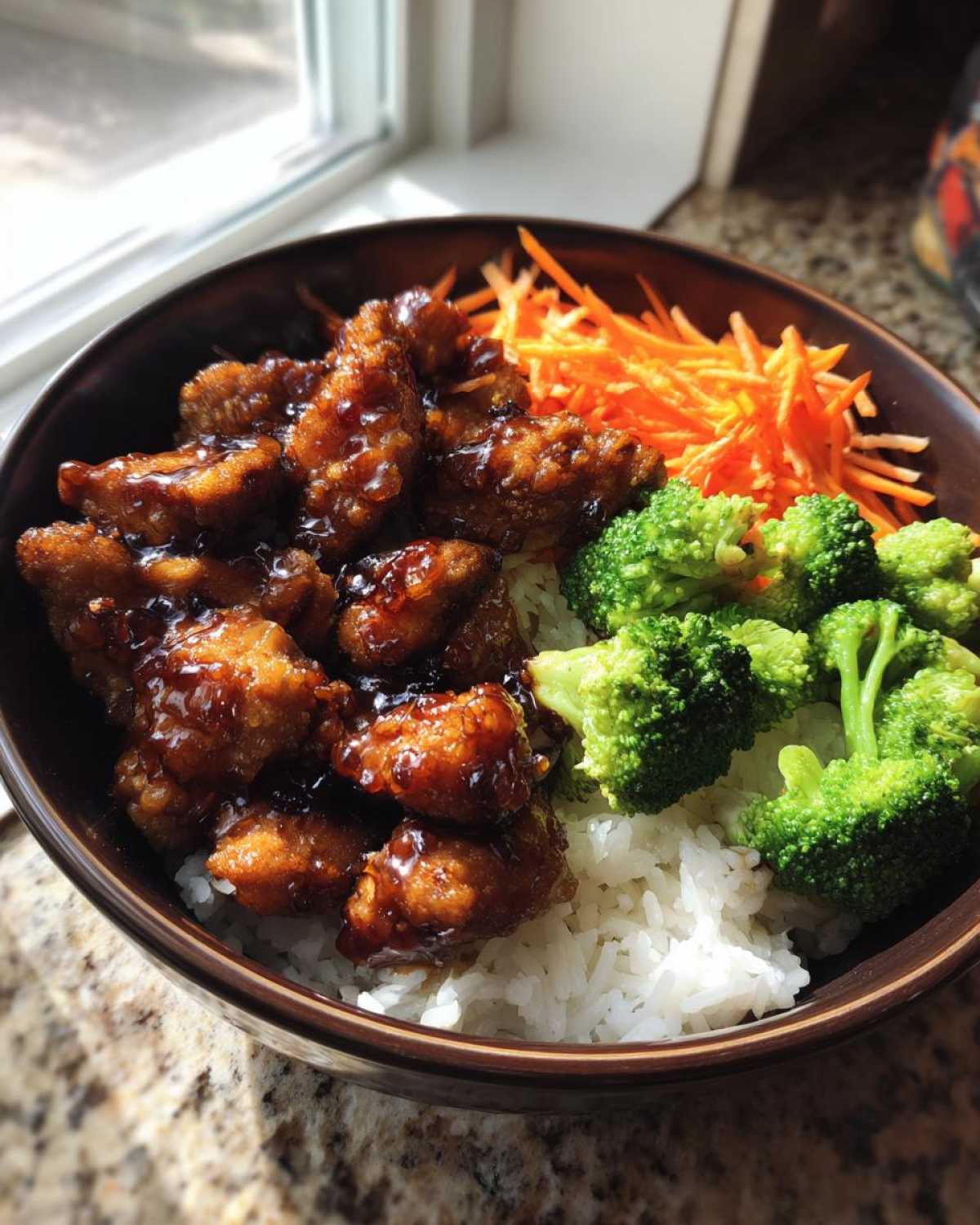 Close-up of a Honey Garlic Chicken Bowl featuring glazed chicken pieces over white rice, served with steamed broccoli and shredded carrots.