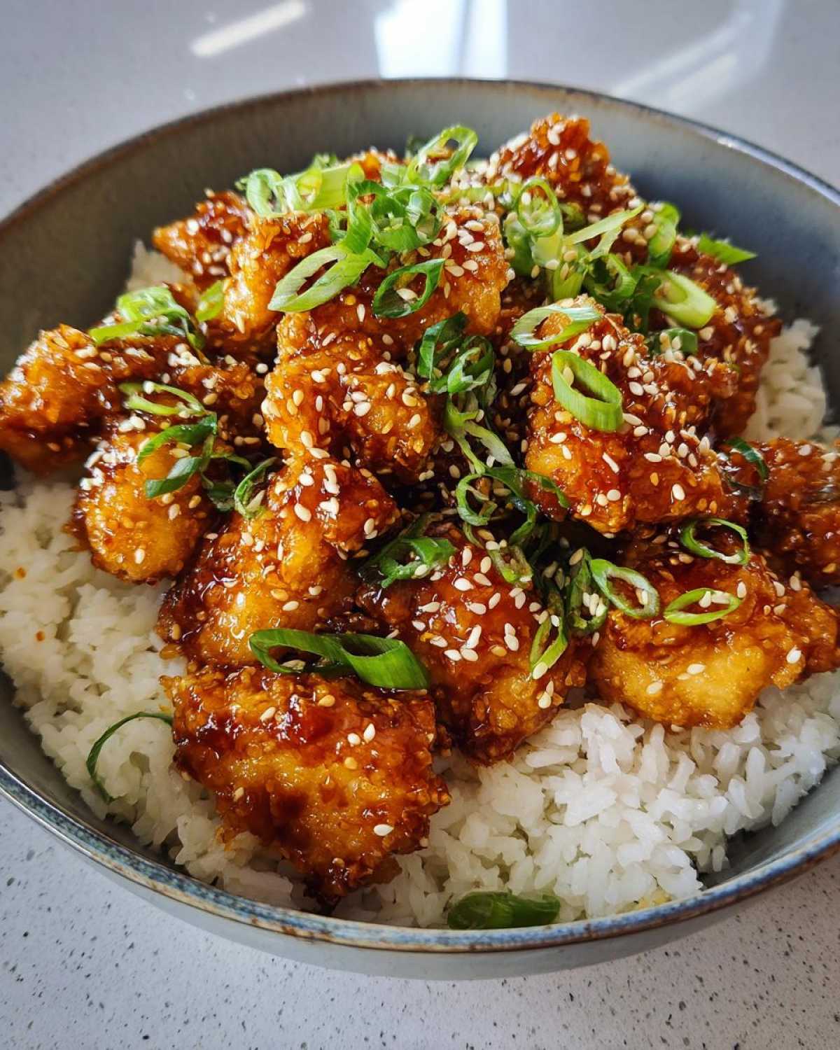 Close-up of crispy chicken pieces coated in honey garlic glaze, served over white rice and topped with sesame seeds and green onions in a bowl.