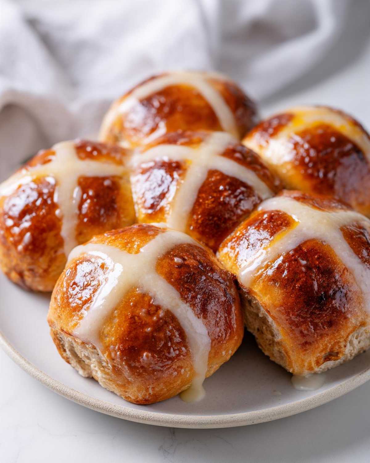 A close-up of freshly baked Hot Cross Buns with Orange Glaze drizzled over the white crosses, resting on a light gray plate.