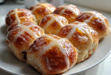 A close-up of freshly baked Hot Cross Buns with Orange Glaze, drizzled with white icing, arranged on a white plate.