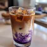 Close-up of a tall glass containing an Iced Lavender Latte with ice, layered milk, espresso, and vibrant purple syrup swirling at the bottom.