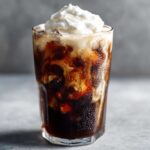 A tall glass of Iced Mocha showing swirls of dark coffee and cream, topped with a generous dollop of whipped cream.
